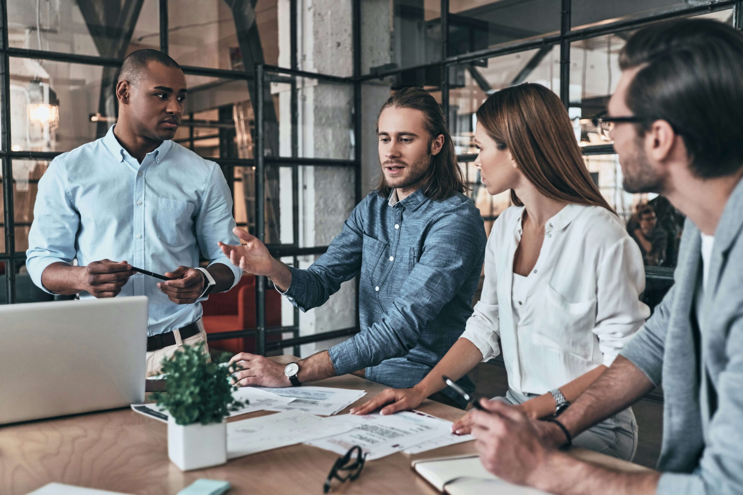 Four professionals collaborating in a modern office meeting around a laptop and strategy documents, demonstrating confidence at work, building self confidence at work through action, ownership of expertise, and aligned team communication.